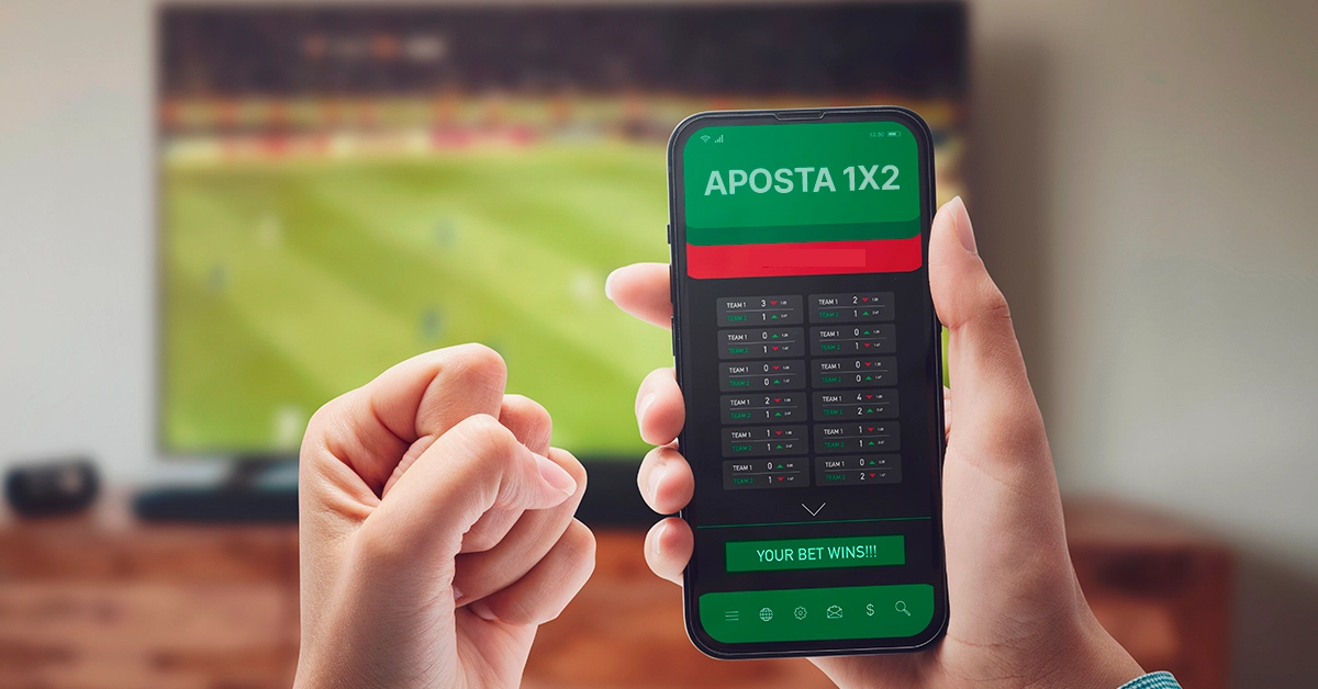 Imagem mostra mãos segurando smartphone aberto em página de apostas. Ao fundo, uma televisão aberta em um jogo de futebol.