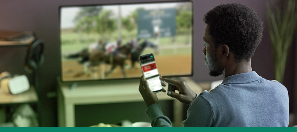 Imagem mostra homem utilizando smartphone aberto em app de aposta enquanto assiste uma corrida de cavalo