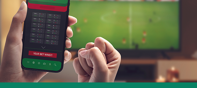 Imagem mostra mão segurando um smartphone aberto em apostas. Ao fundo, uma televisão transmitindo uma partida de futebol.