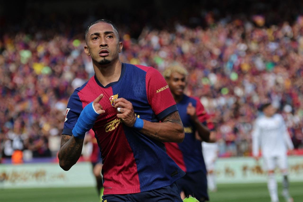 Raphinha apontando para escudo do Barcelona (foto: Lluis Gene / AFP)